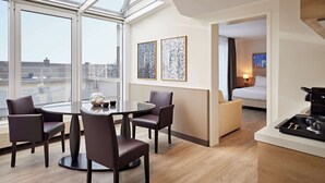 Ein modernes Hotelzimmer mit einem großen Oberlicht, einem Essbereich mit rundem Tisch und vier Stühlen, einem Bett mit Nachttisch sowie zwei gerahmten abstrakten Kunstwerken an der Wand.