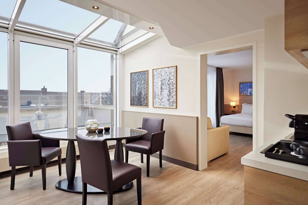 Une chambre d’hôtel moderne dotée d’une grande verrière, d’un coin repas avec une table ronde et quatre chaises, d’un lit avec une table de chevet, et de deux œuvres d’art abstraites encadrées accrochées au mur.