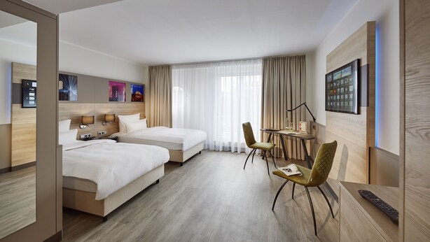 Ein Hotelzimmer mit zwei Betten, einem Schreibtisch mit Stuhl, einem Fernseher und einem Fenster mit Vorhängen.