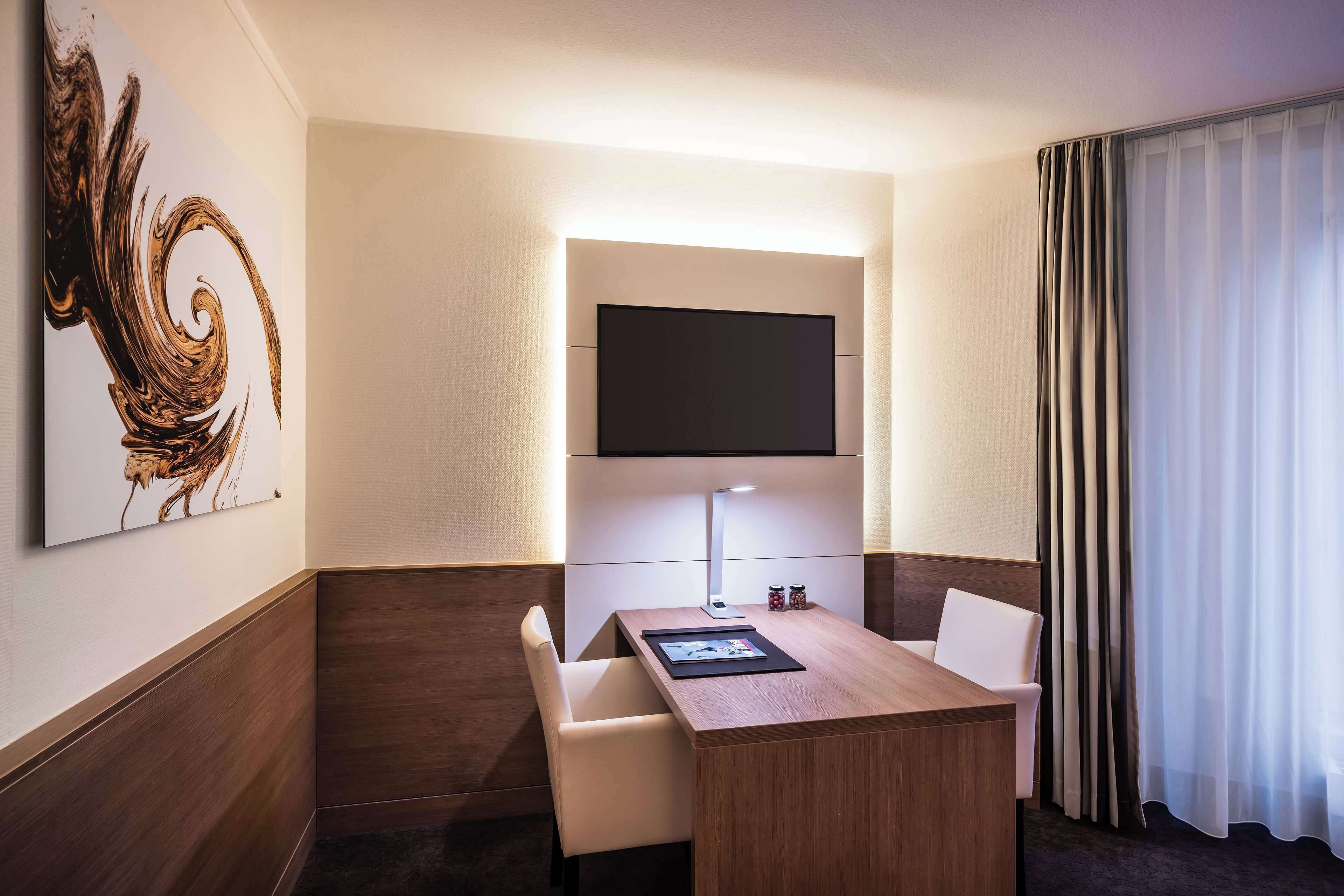 A hotel room with a desk, two chairs, a flat-screen TV mounted on the wall, a lamp, and a large window with curtains.
