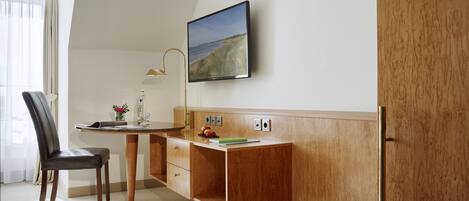 Una habitación de hotel moderna con un escritorio de madera, una silla, un televisor de pantalla plana montado en la pared y una ventana con cortinas translúcidas.