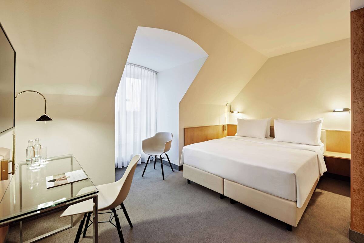 Une chambre d’hôtel avec un grand lit, un bureau avec une chaise, une table en verre et une lampe moderne.
