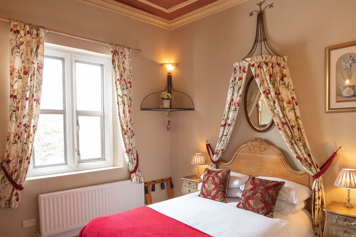 A bedroom with a large bed, a window with curtains, a radiator, a bedside table with a lamp, and a tray with cups.