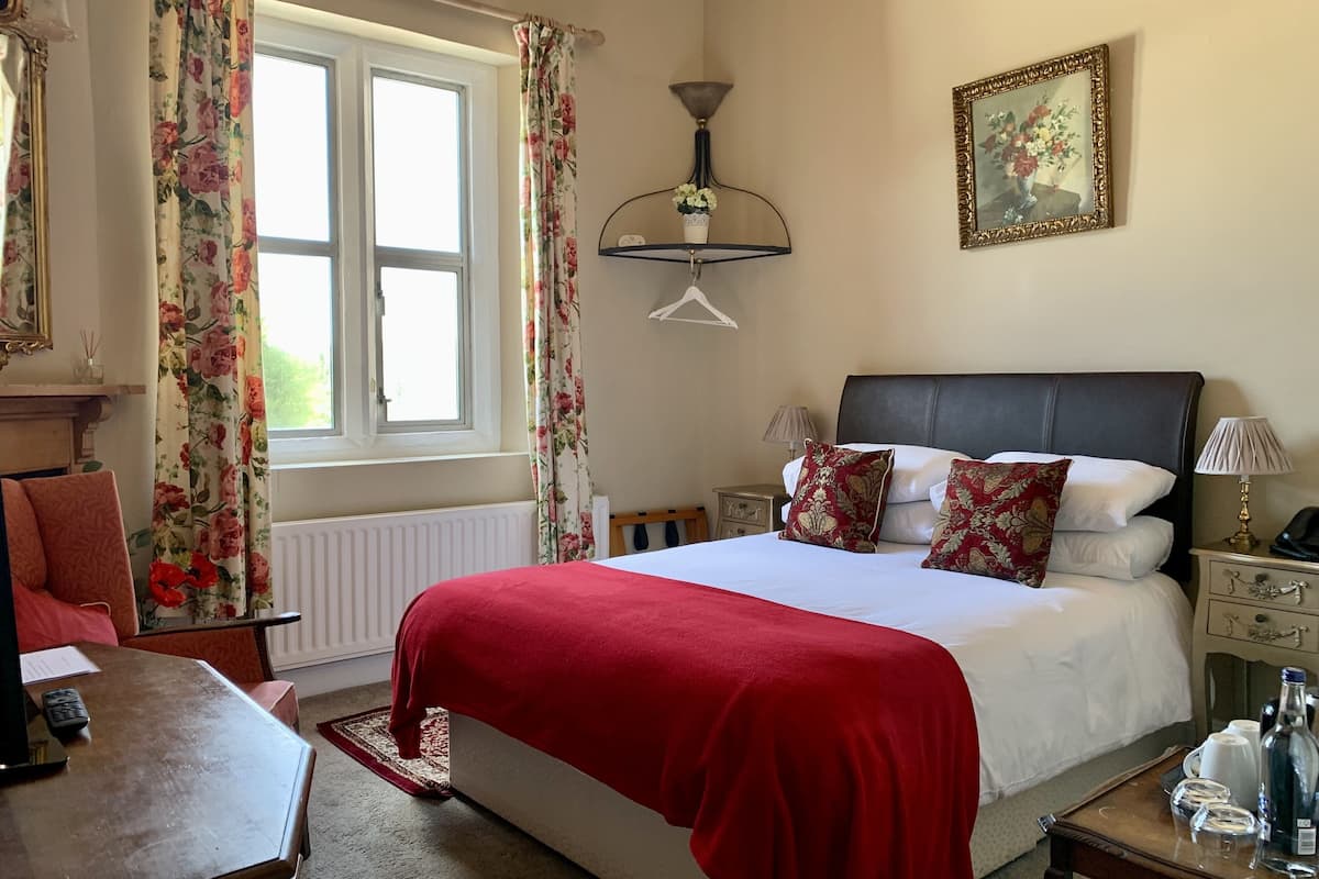 A hotel room with a bed, bedside tables, a desk, a chair, a window with curtains, a wall-mounted lamp, and a framed picture.