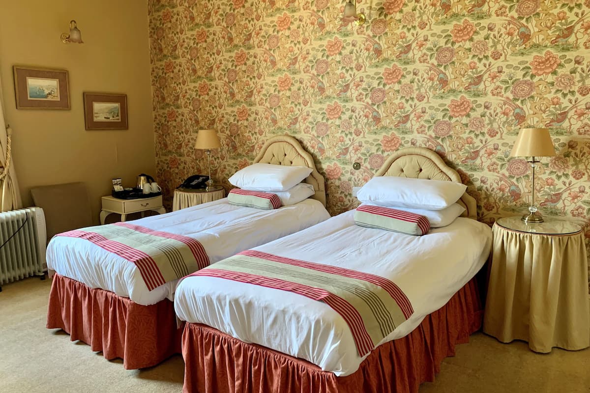 A hotel room with two beds, floral wallpaper, a radiator, bedside tables with lamps, and framed pictures on the wall.