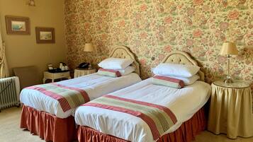 Habitación de hotel con dos camas, papel tapiz floral, un radiador, mesitas de noche con lámparas y cuadros en la pared.