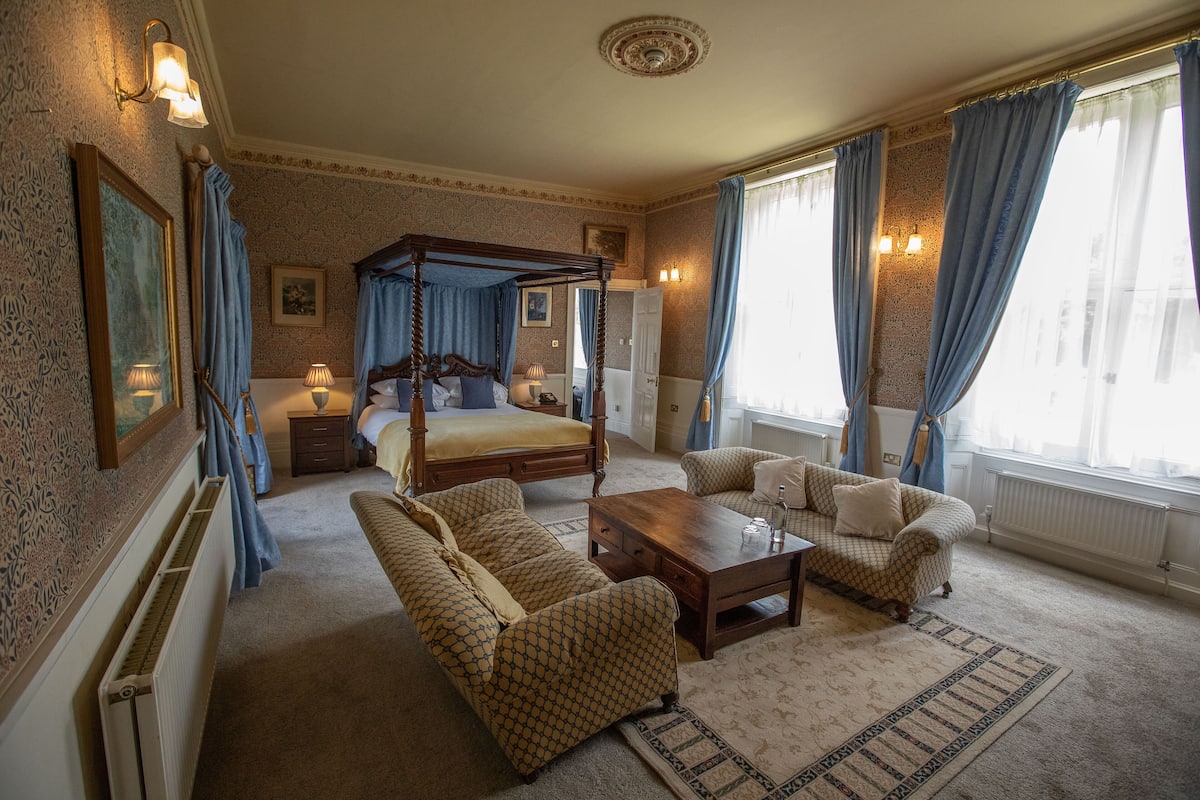A bedroom with a four-poster bed, a sitting area with a sofa and armchairs, a wooden coffee table, and a large window with curtains.
