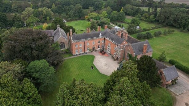 A large, historic building with a symmetrical facade, surrounded by lush greenery and open fields. - Grafton Manor (Bromsgrove)