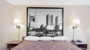 A hotel room with a bed, two bedside tables, a cityscape artwork on the wall, and two lamps.