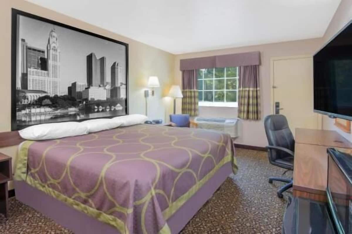 A hotel room with a bed, a desk, a chair, a television, a window with curtains, and a cityscape painting on the wall.