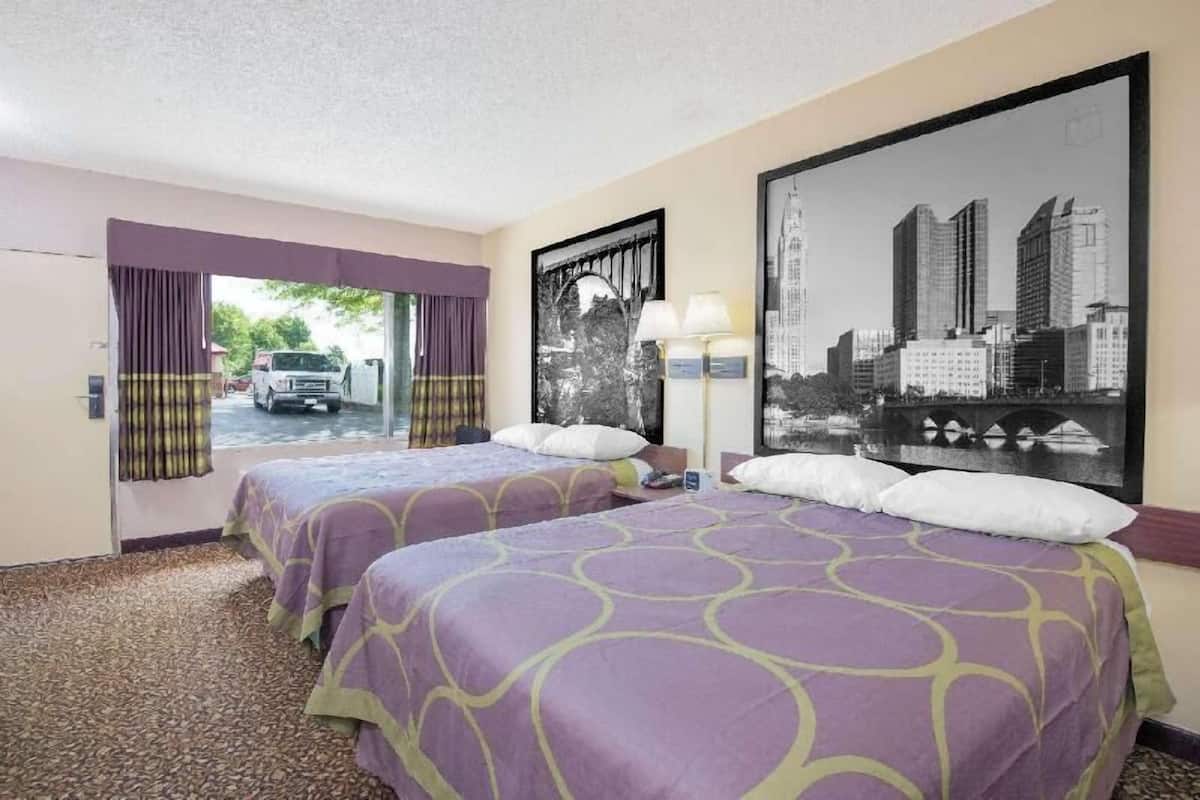 A hotel room with two beds, a cityscape painting, a window with curtains, and a lamp.