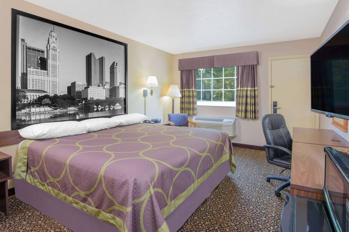 Een hotelkamer met een groot bed, een bureau, een stoel, een televisie, een raam met gordijnen en een stadsgezichtschilderij aan de muur.