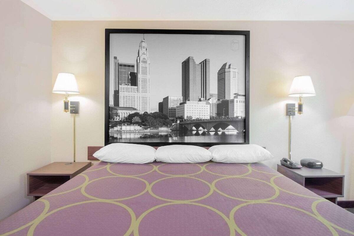 A hotel room with a bed, two bedside tables, a cityscape painting, and two wall-mounted lamps.