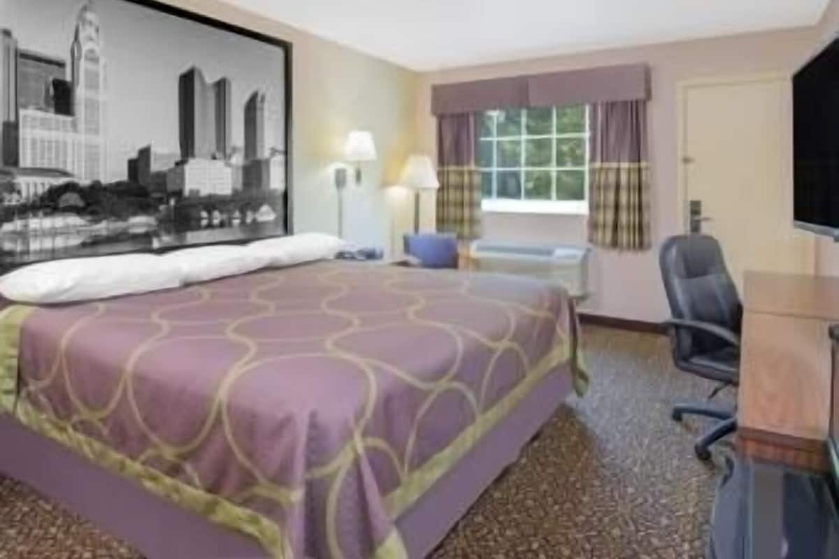 A hotel room with a bed, a desk, a chair, a window with curtains, and a cityscape painting on the wall.