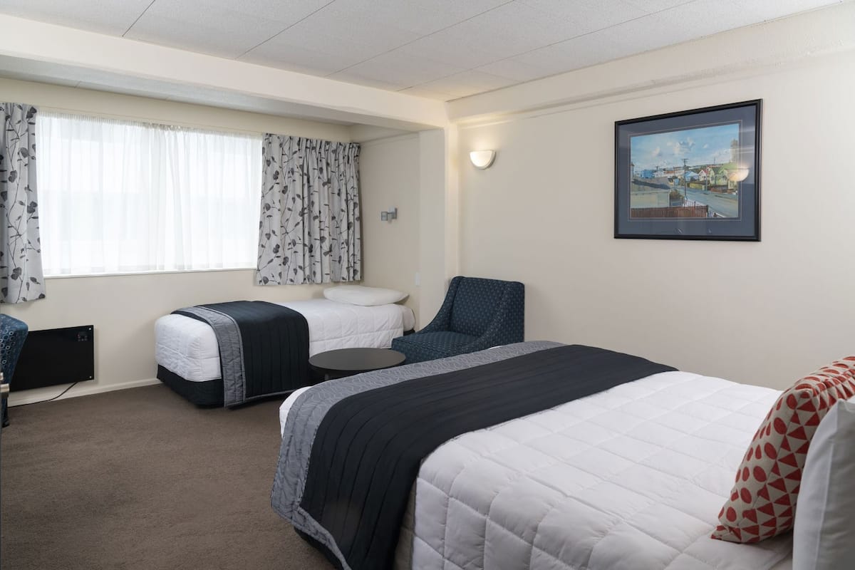 Ein Hotelzimmer mit zwei Betten, einem Sessel, einem Schreibtisch, einem Fenster mit Vorhängen und einem gerahmten Bild an der Wand.