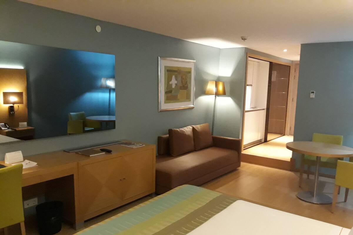 A hotel room with a bed, a desk, a chair, a sofa, a dining table, and a mirror.
