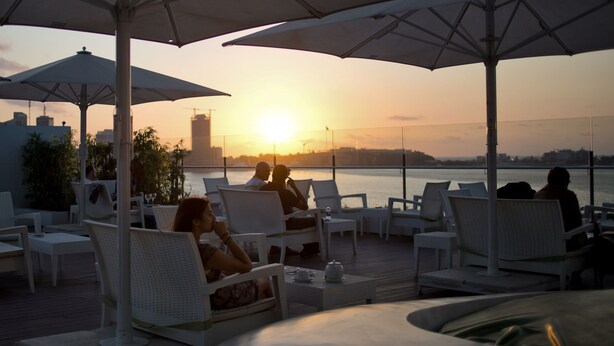 Une terrasse sur le toit, avec des chaises en osier blanc et des parasols, d’où l’on peut admirer un coucher de soleil sur une étendue d’eau.