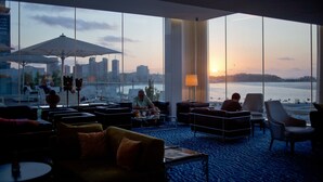 Un espace de détente moderne doté de grandes fenêtres offrant une vue sur les grat-ciel d’une ville et sur une étendue d’eau au coucher du soleil.