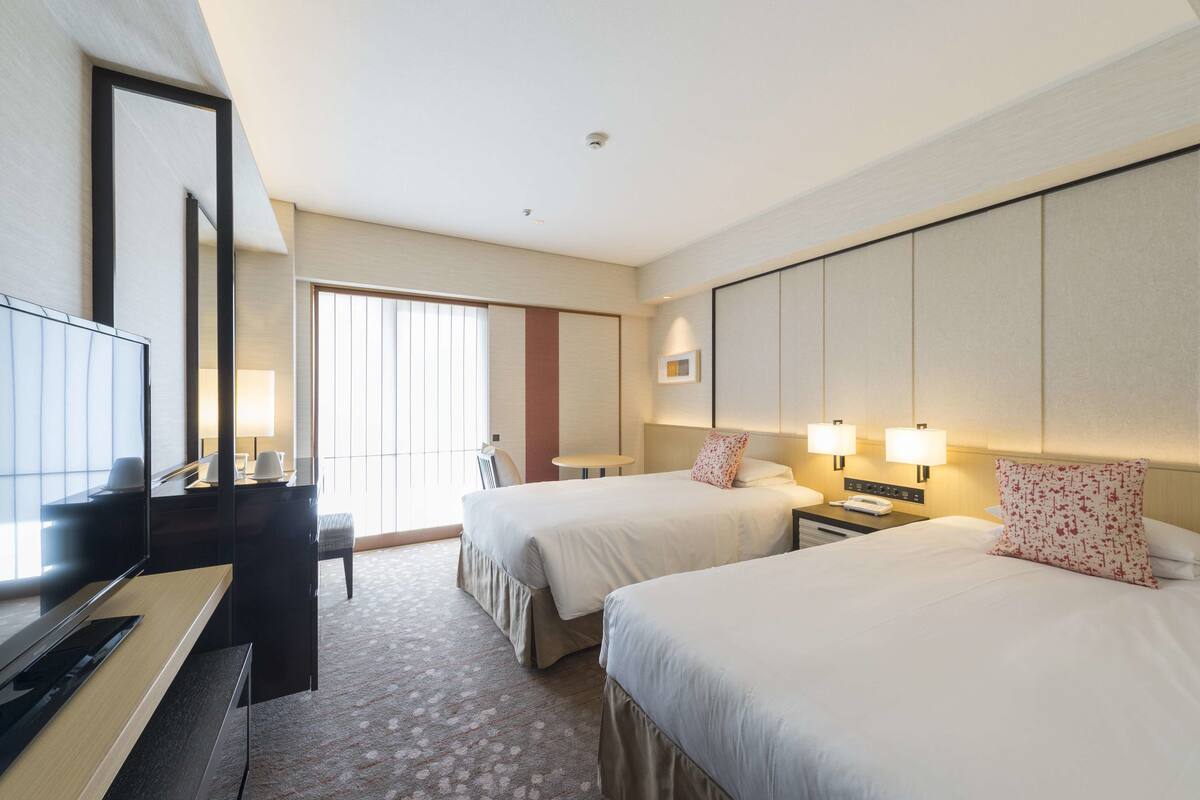 Hotellihuone, jossa on kaksi sänkyä, työpöytä, tuoli, televisio ja suuri ikkuna.