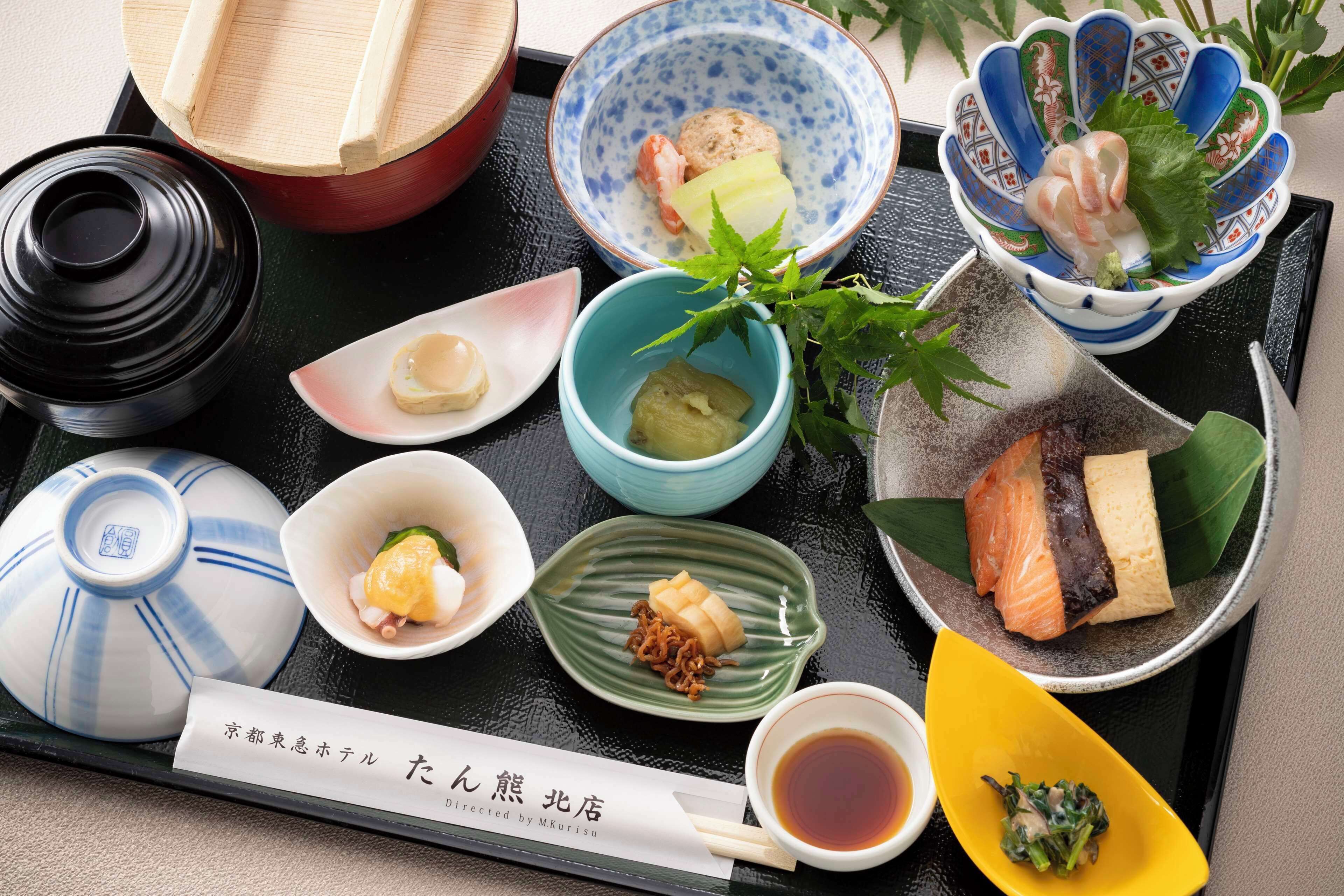 A variety of Japanese dishes arranged on a black tray, including sushi, sashimi, and other traditional foods, with a small bowl of soy sauce and a nameplate indicating the restaurant.