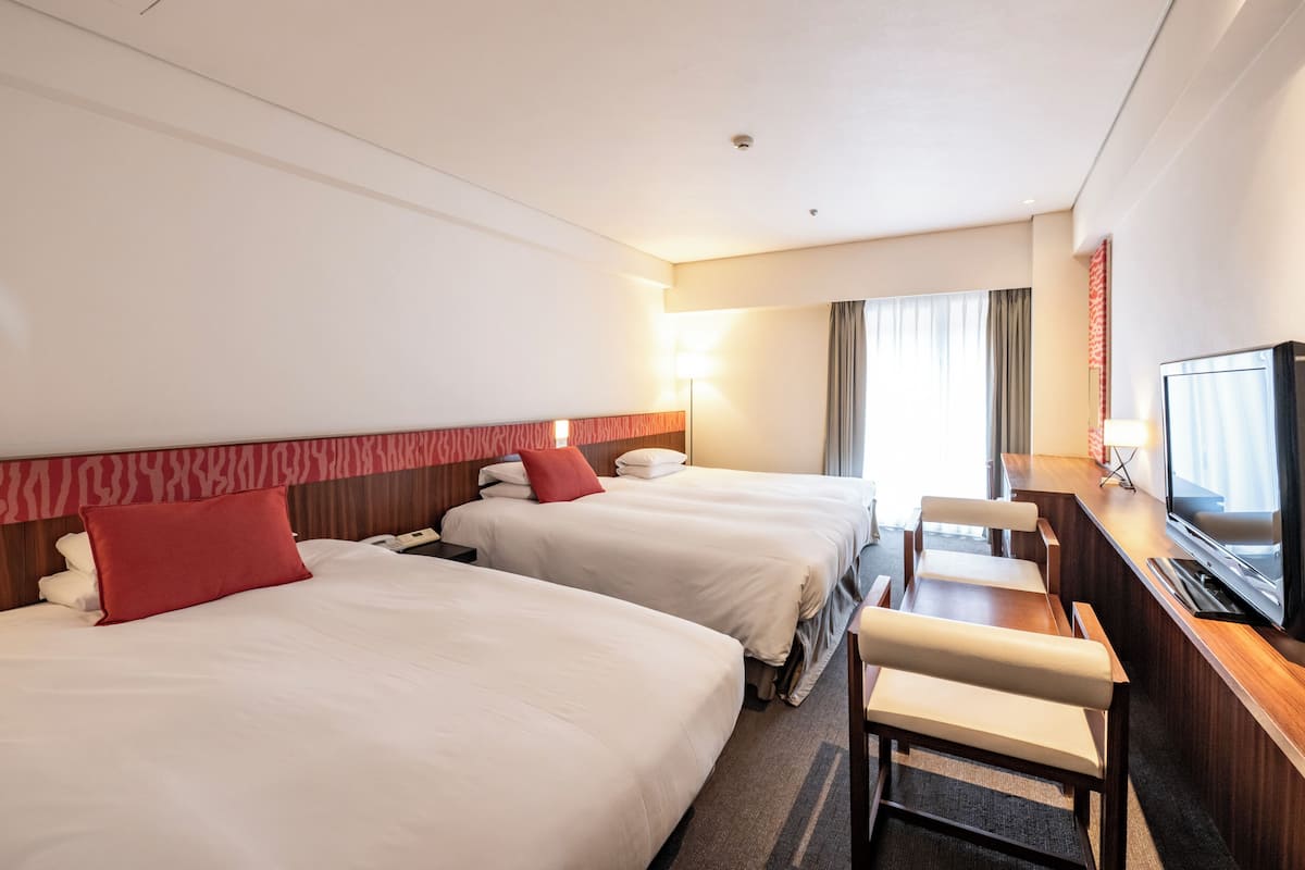 A hotel room with two beds, a desk with a TV, and a window with curtains.