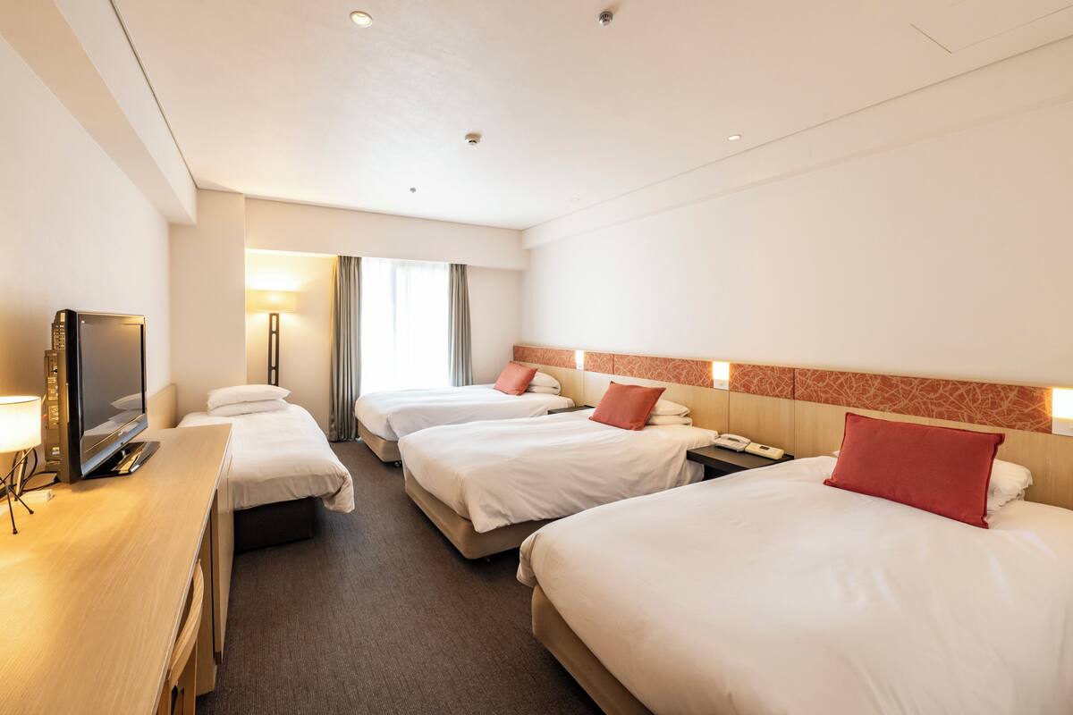 Hotellihuone, jossa on kolme sänkyä, työpöytä, jossa on televisio, ja lamppu.