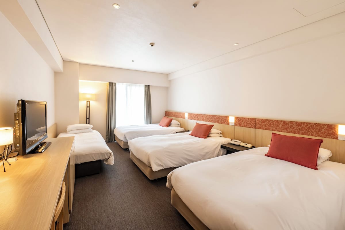 A hotel room with three beds, a desk with a TV, and a lamp.