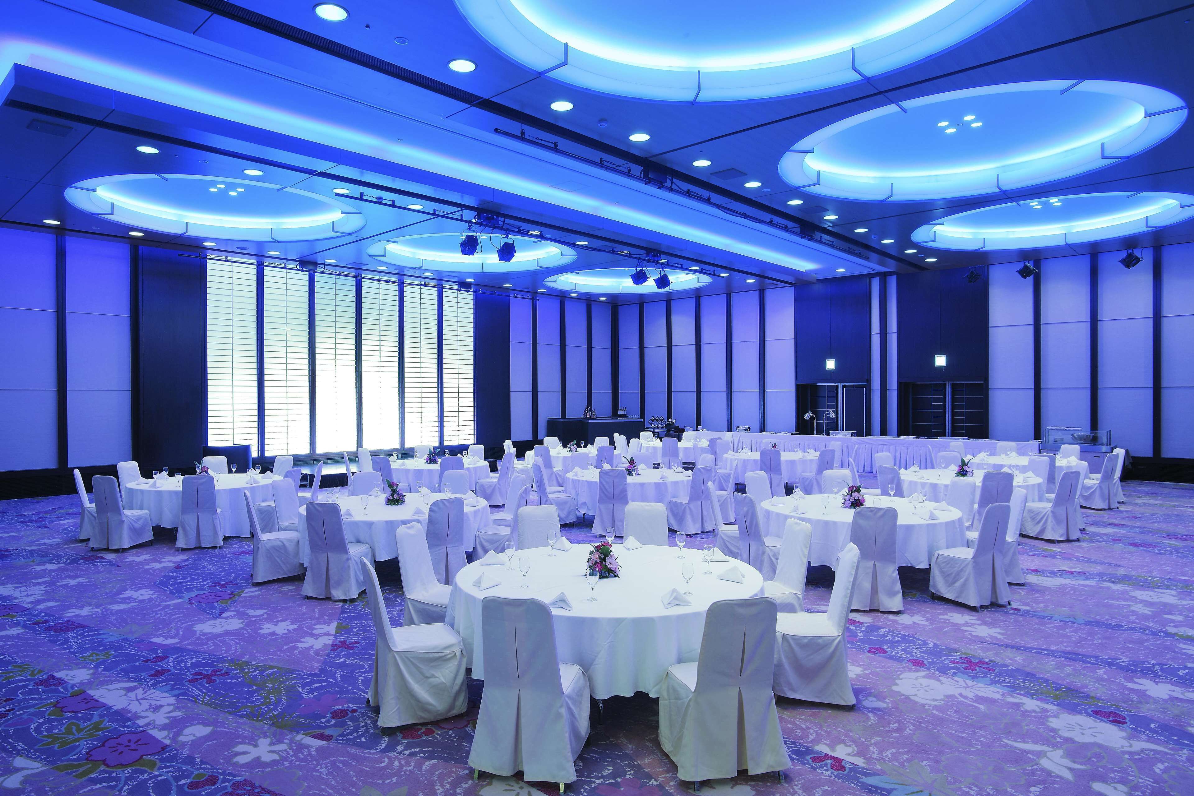 A large banquet hall with round tables and white chairs, featuring a blue-lit ceiling and a floral carpet.