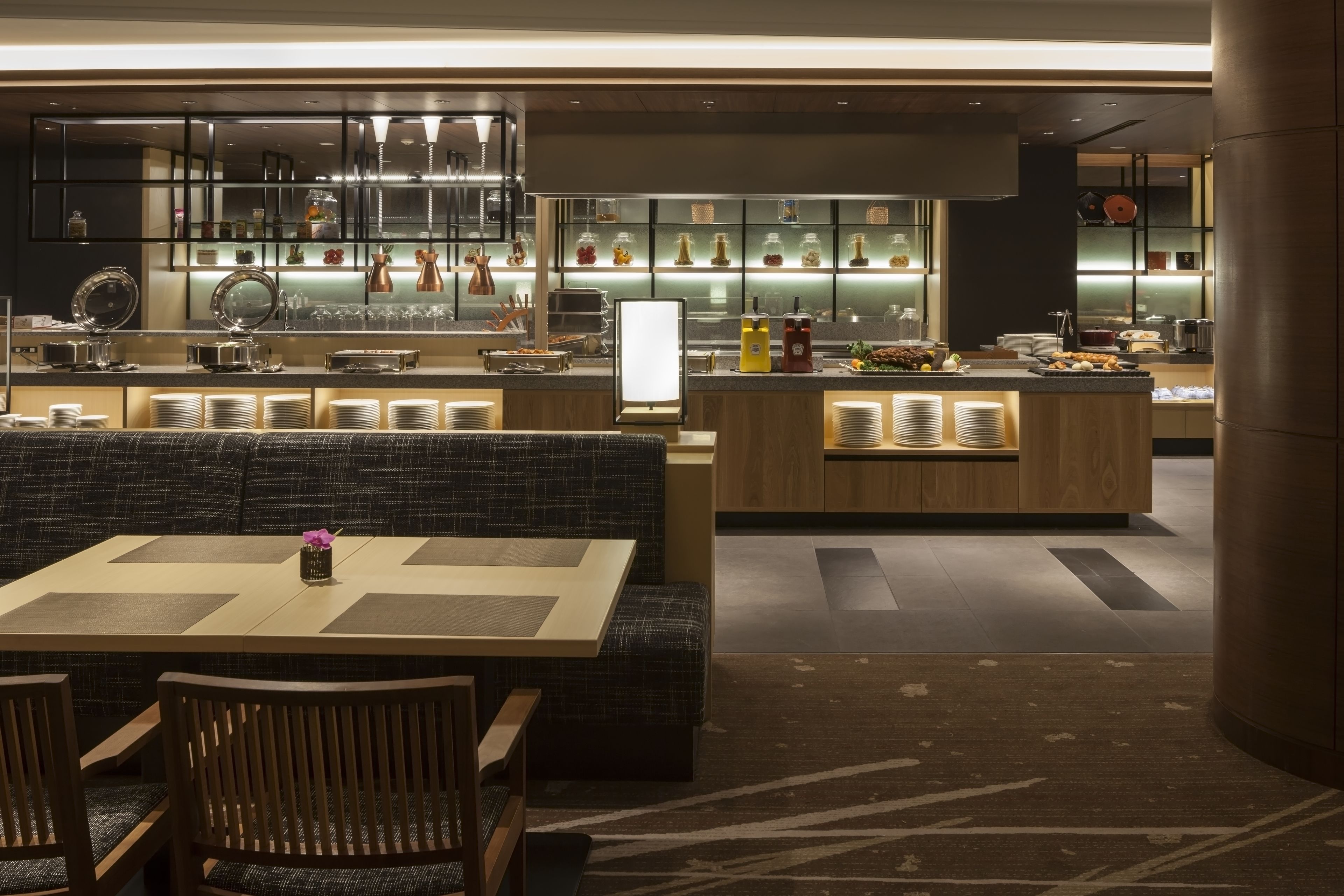 A modern restaurant with a central dining area featuring a wooden table and cushioned seating. There is a bar area with a counter and various food items displayed.