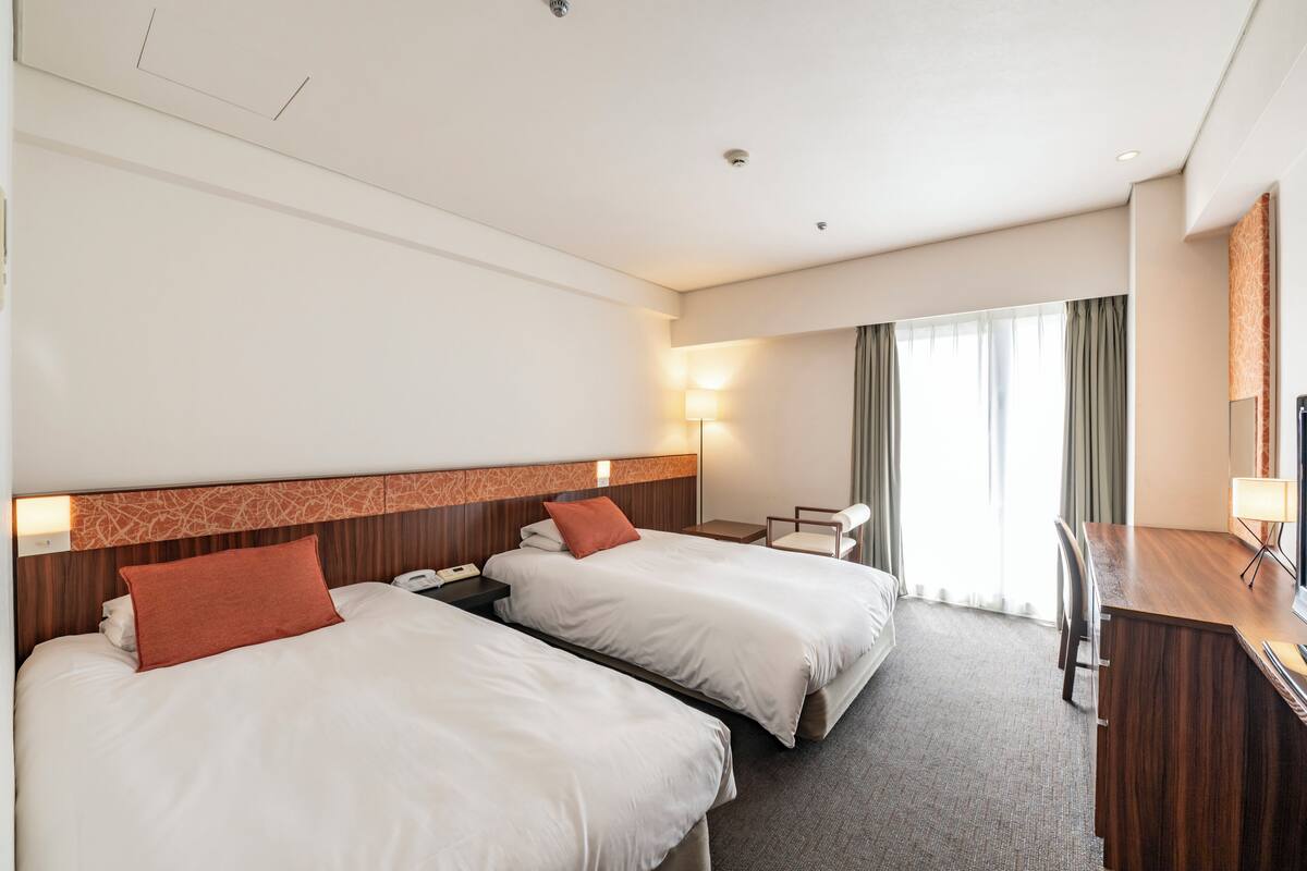 Hotellihuone, jossa on kaksi sänkyä, työpöytä, tuoli, lamppu ja suuri ikkuna, jossa on verhot.