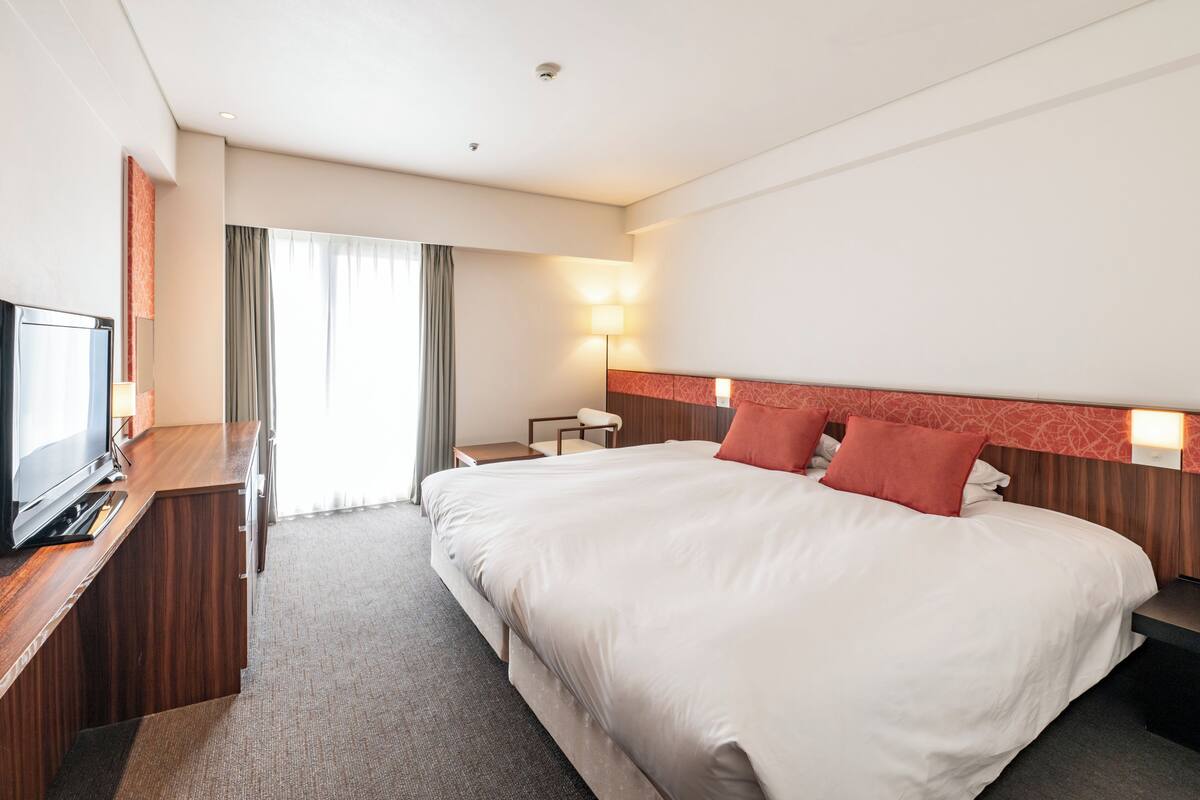 Hotellihuone, jossa on sänky, televisio, työpöytä, lamppu ja ikkuna, jossa on verhot.