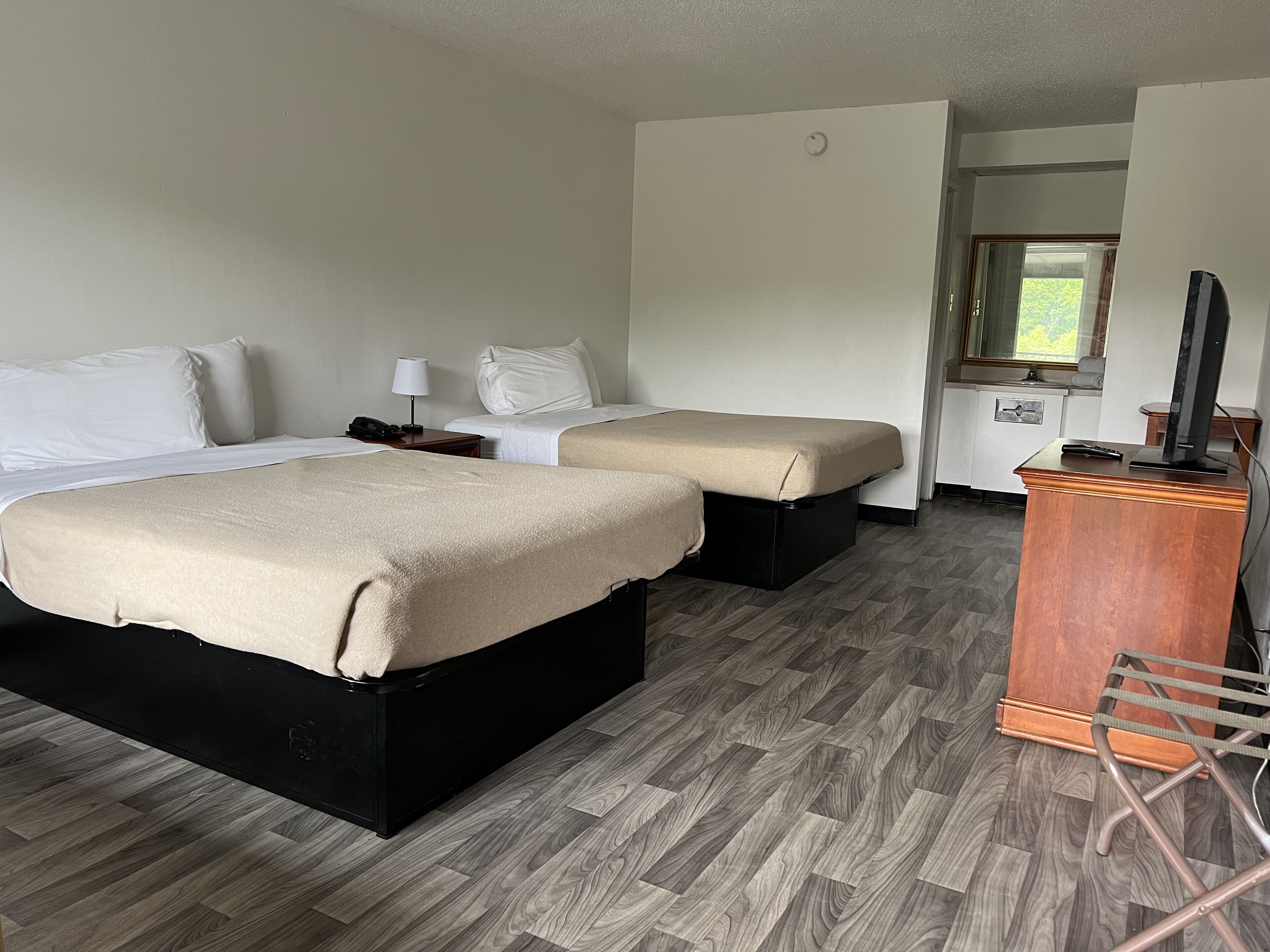Ein Hotelzimmer mit zwei Betten, Holzboden, Schreibtisch mit Fernseher und Fenster mit Blick auf Grünfläche.