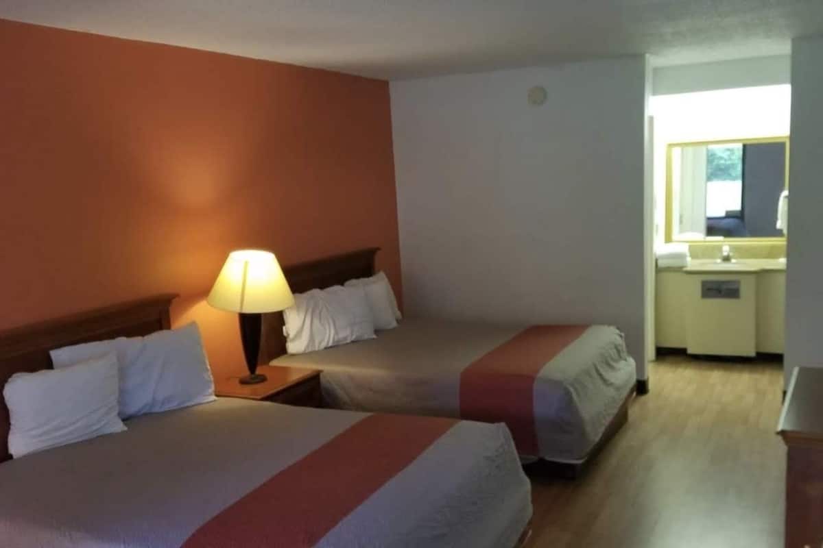 A hotel room with two beds, a nightstand with a lamp, a small desk with a computer, and a bathroom visible through an open door.
