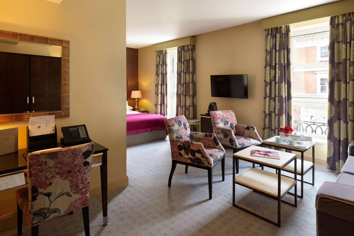 A hotel room with a bed, a TV, floral armchairs, a small table, and a sofa.