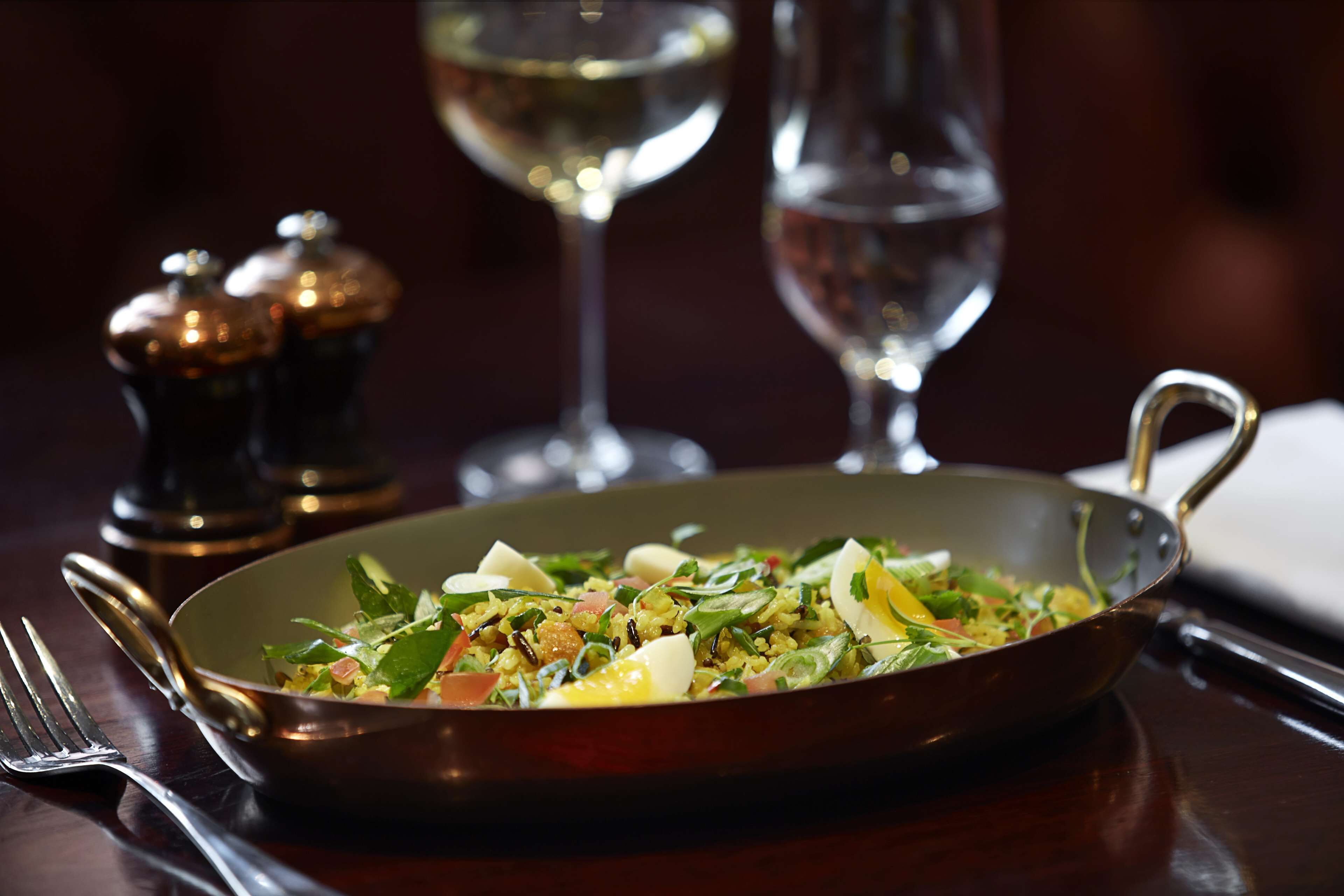 A bowl of salad on a table with a fork and a wine glass in the background.