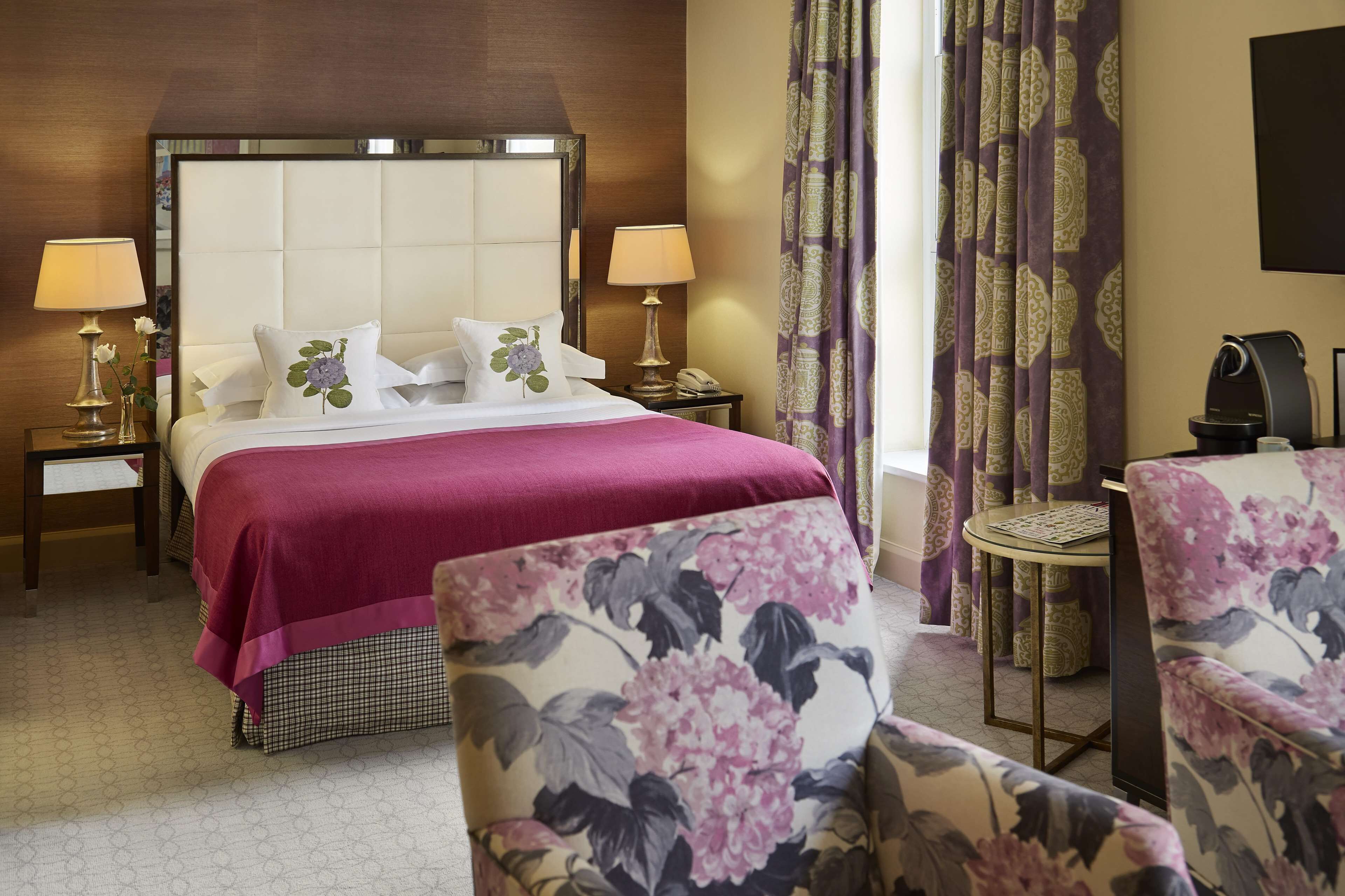 Une chambre d’hôtel avec un grand lit, deux lampes de chevet, une petite table ronde et des fauteuils à motifs floraux.