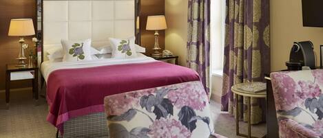 Une chambre d’hôtel avec un grand lit, deux lampes de chevet, une petite table ronde et des fauteuils à motifs floraux.