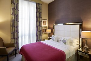 Una camera d'albergo con un letto grande, una sedia, un comodino con lampade e una finestra con tende.