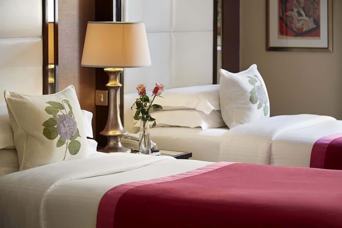 A hotel room with two beds, a bedside table with a lamp, and a vase with flowers.