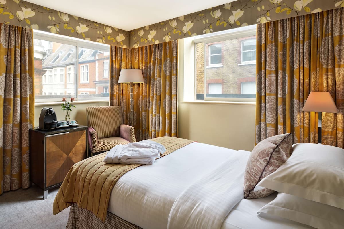 A hotel room with a bed, bedside table, lamp, chair, and a view of buildings through the window.