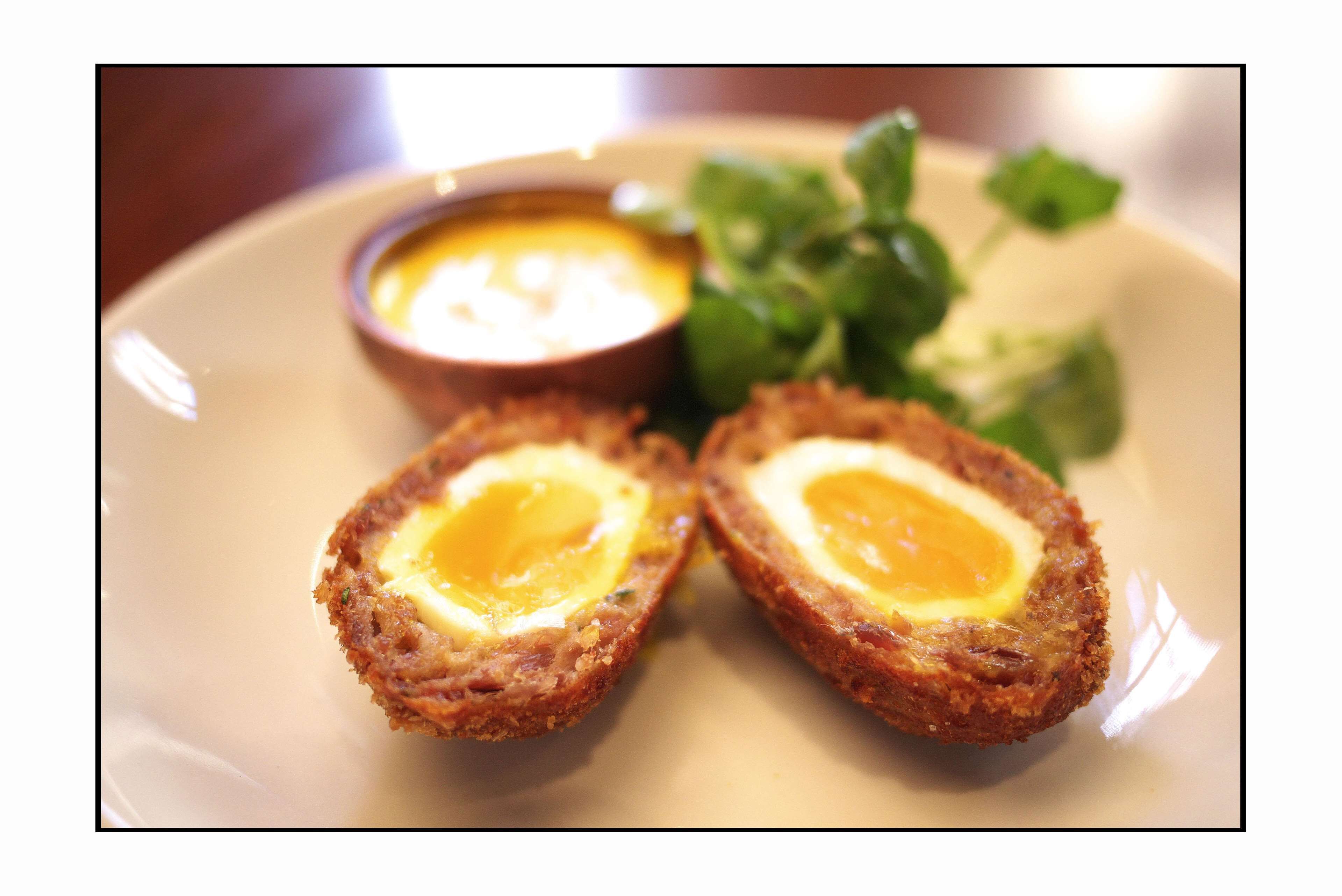 A plate with two Scotch eggs and a small bowl of sauce.