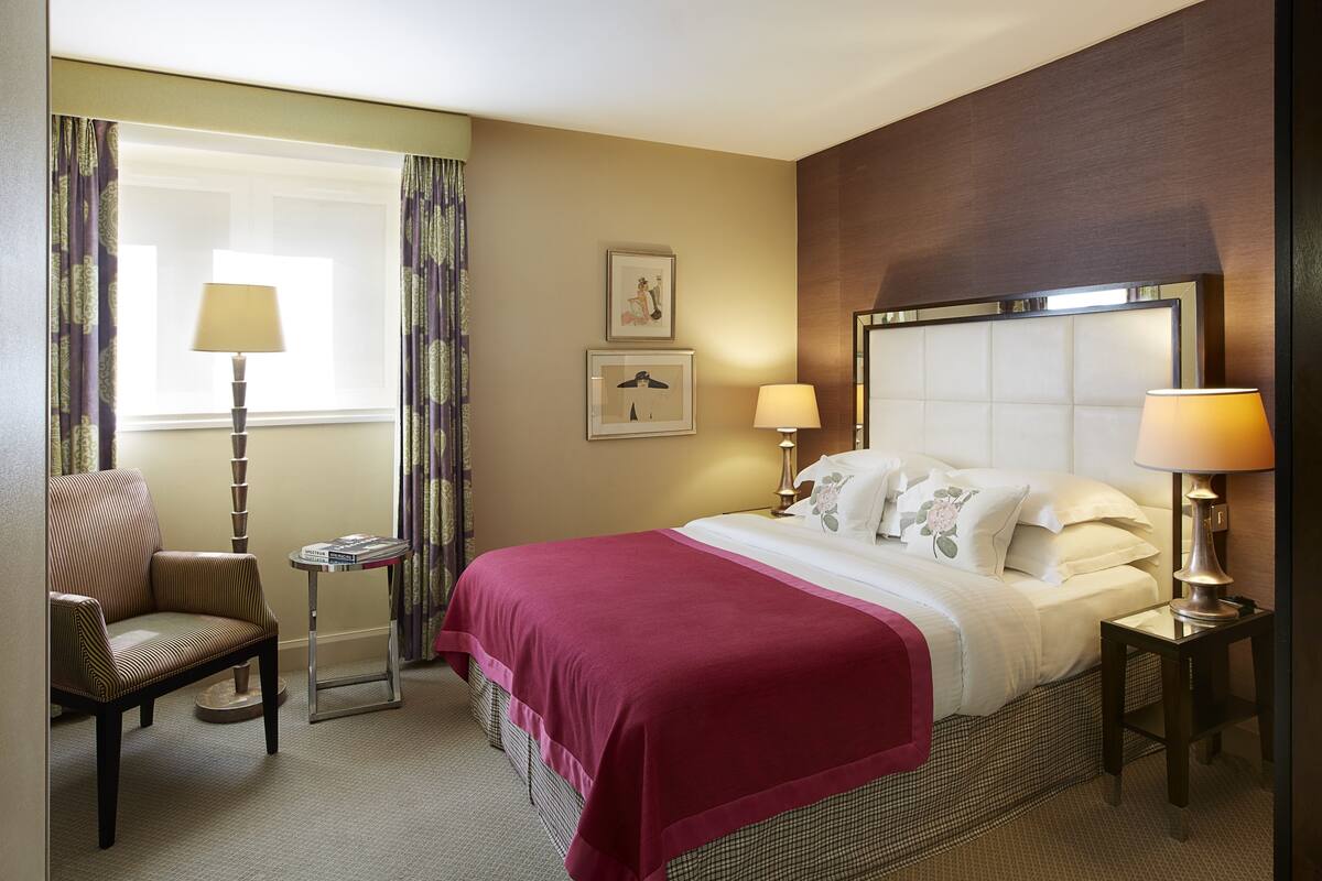 A hotel room with a large bed, two bedside tables, a chair, a window with curtains, and two framed pictures on the wall.
