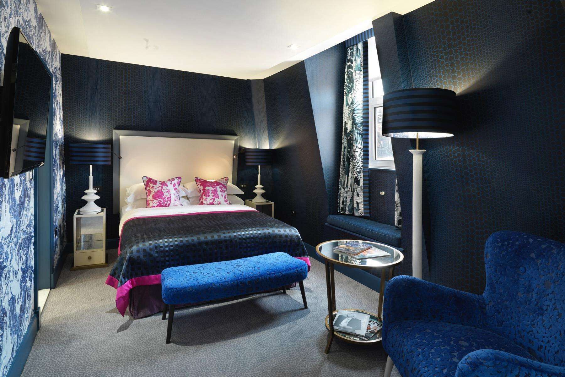Habitación de hotel moderna con una cama grande, un sillón azul, una mesita redonda y una ventana con cortinas estampadas.