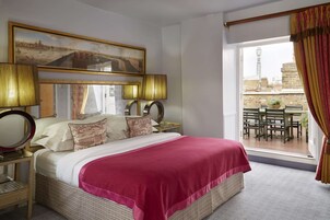 Ein Hotelzimmer mit einem großen Bett, zwei Nachttischen mit Lampen, Blick auf die Stadt durch ein Fenster und einem Balkon mit Gartenmöbeln.