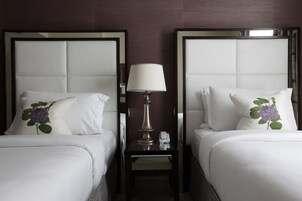 A hotel room with two beds, each with a white headboard and a pillow with a purple flower design. There is a nightstand between the beds with a lamp and a phone.