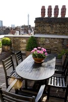 Ein runder Holztisch mit sechs Stühlen, umgeben von Topfpflanzen, befindet sich auf einer Dachterrasse mit einer Ziegelmauer und Stadtansicht im Hintergrund.