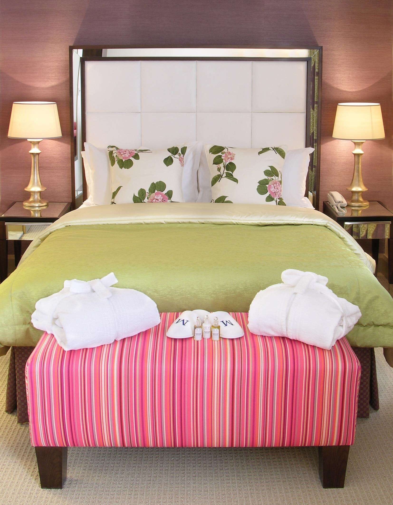 A neatly made bed with floral pillows, a green bedspread, and a striped ottoman with towels and toiletries.