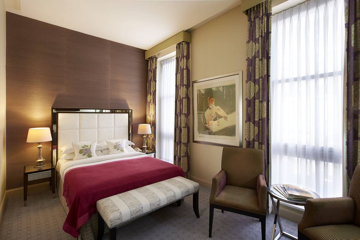 A hotel room with a large bed, two bedside lamps, a bench, two armchairs, a small table, and a framed picture on the wall.
