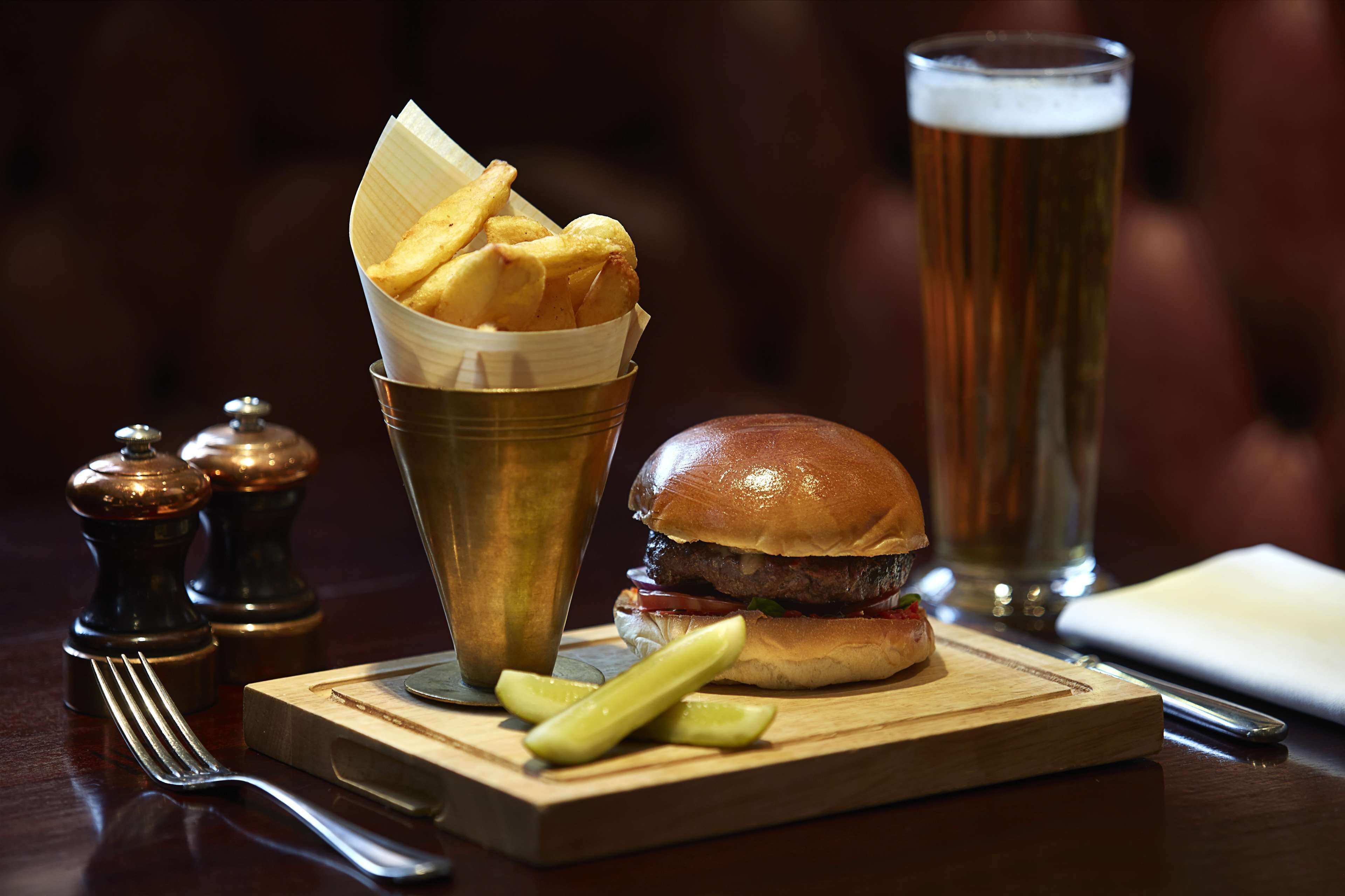 A meal with a burger and fries on a wooden board, a glass of beer, and condiments on a table.