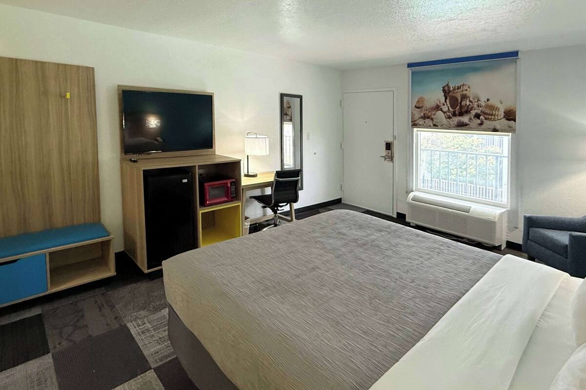 A hotel room with a large bed, a desk with a chair, a television, a wardrobe, and a window with a view of the beach.